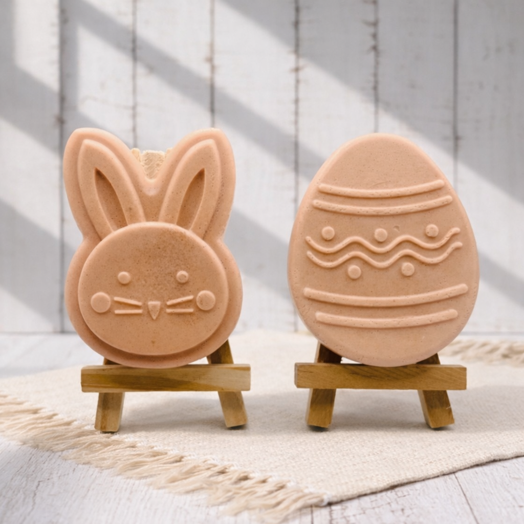 handmade vegan soap gift set featuring a cute bunny face and a decorated Easter egg with wave patterns, displayed on wooden easels against a rustic white farmhouse decor background with spring flowers.