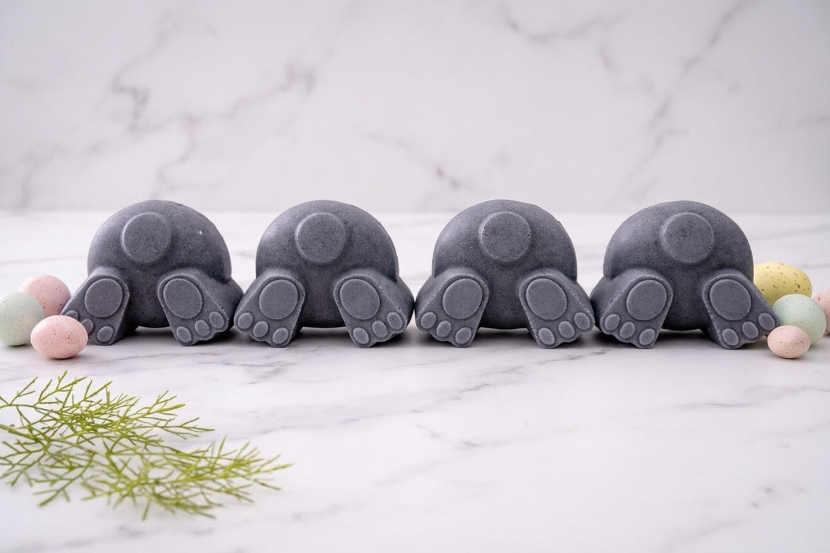 Row of four charcoal gray activated charcoal vegan soaps shaped as bunny bums and tails on white marble. The natural skincare set includes decorative green sprigs and colourful miniature speckled eggs.