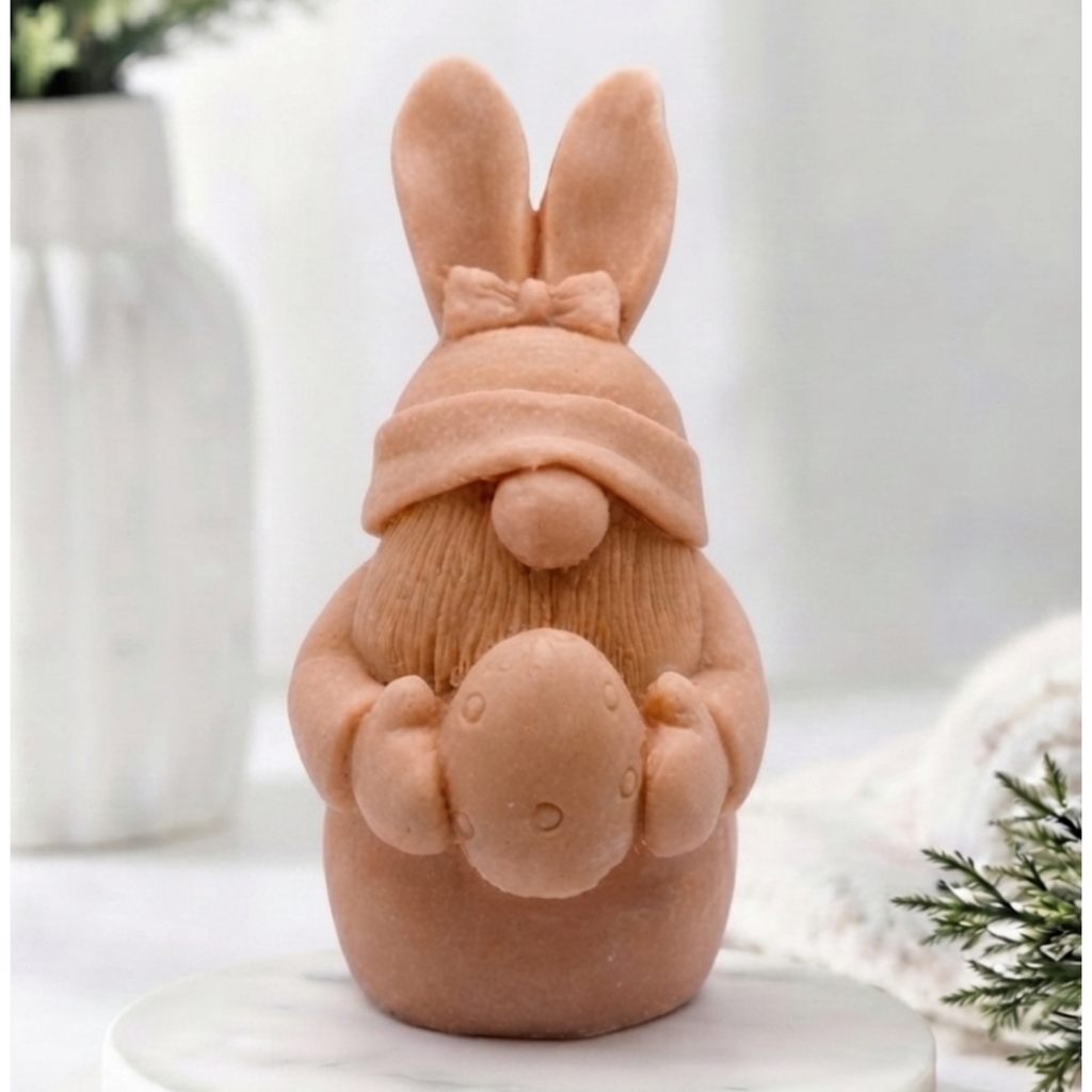 A detailed handmade vegan soap in a natural colour, sculpted as a whimsical Easter gnome (gonk) with long rabbit ears and a bow on its hat, holding a patterned Easter egg, displayed on a light wooden surface. Easter gift 