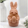 A detailed handmade vegan soap in a natural colour, sculpted as a whimsical Easter gnome (gonk) with long rabbit ears and a bow on its hat, holding a patterned Easter egg, displayed on a light wooden surface. Easter gift 