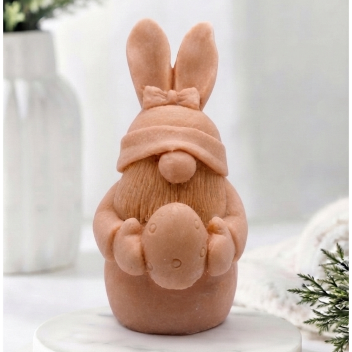 A detailed handmade vegan soap in a natural colour, sculpted as a whimsical Easter gnome (gonk) with long rabbit ears and a bow on its hat, holding a patterned Easter egg, displayed on a light wooden surface. Easter gift 