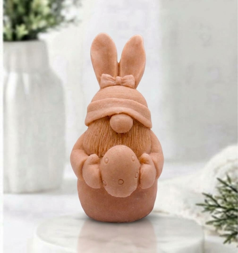 A detailed terracotta coloured handmade vegan soap in a natural colour, sculpted as a whimsical Easter gnome (gonk) with long rabbit ears and a bow on its hat, holding a patterned Easter egg, displayed on a light wooden surface. Easter gift 