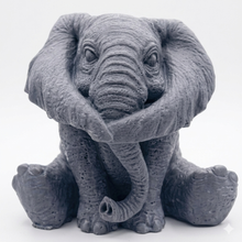 Load image into Gallery viewer, A crisp product photograph of a detailed, grey artisan bar of soap intricately molded into the shape of a sitting baby elephant. The elephant is depicted holding its trunk with its front legs crossed in a "hear no evil" or hugging pose, showcasing realistic skin textures, expressive eyes, and large ears. The product is perfectly centered against a seamless, solid, plain white background.
