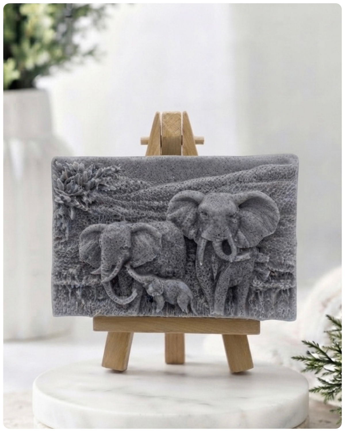A rectangular handmade vegan soap in a warm yellow tone, featuring a detailed relief carving of an elephant family—two adults and a small calf—walking through a textured savannah landscape, displayed on a miniature wooden easel.