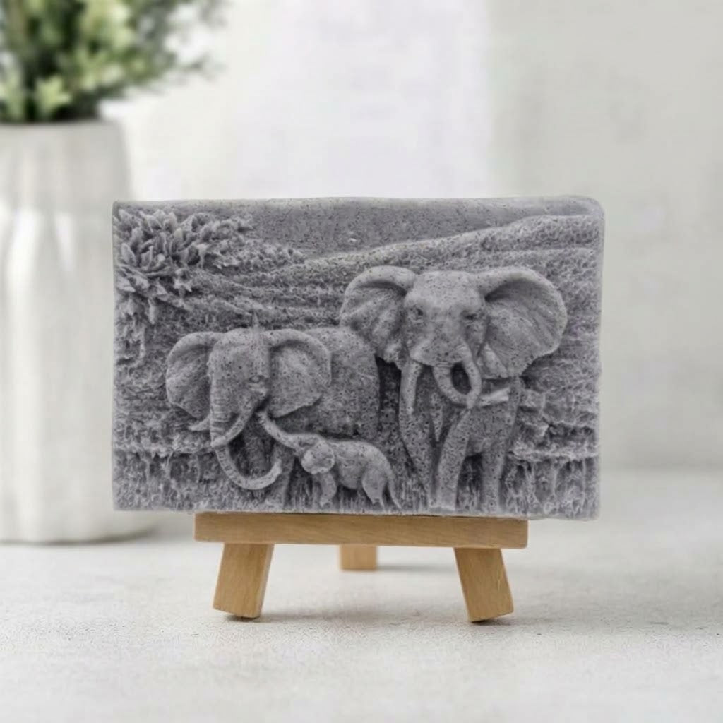A grey, highly detailed relief-carved soap slab showing a family of three elephants – two larger and one smaller – within a carved landscape, presented head-on while supported by a small wooden easel.
