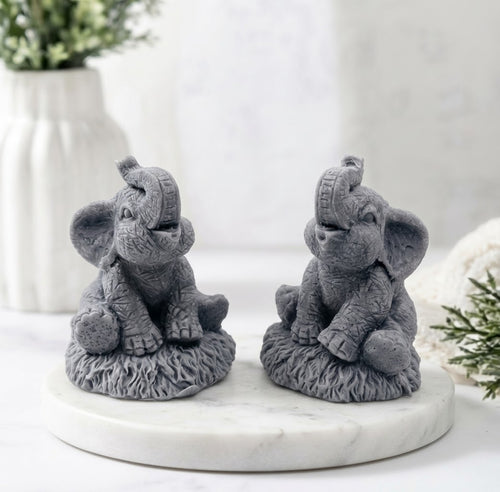 Two gray, detailed elephant-shaped vegan soap figurines, with trunks raised, sit on a white marble trivet.