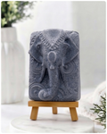 A rectangular handmade vegan soap in a speckled slate-grey finish, featuring a highly detailed relief of an elephant's head adorned with ornate, traditional patterns and a decorative floral background, displayed on a miniature wooden easel.