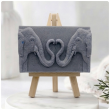 Load image into Gallery viewer, A rectangular handmade vegan soap  featuring a detailed relief of two elephants facing each other with their trunks entwined to form a heart shape, displayed on a miniature wooden easel.
