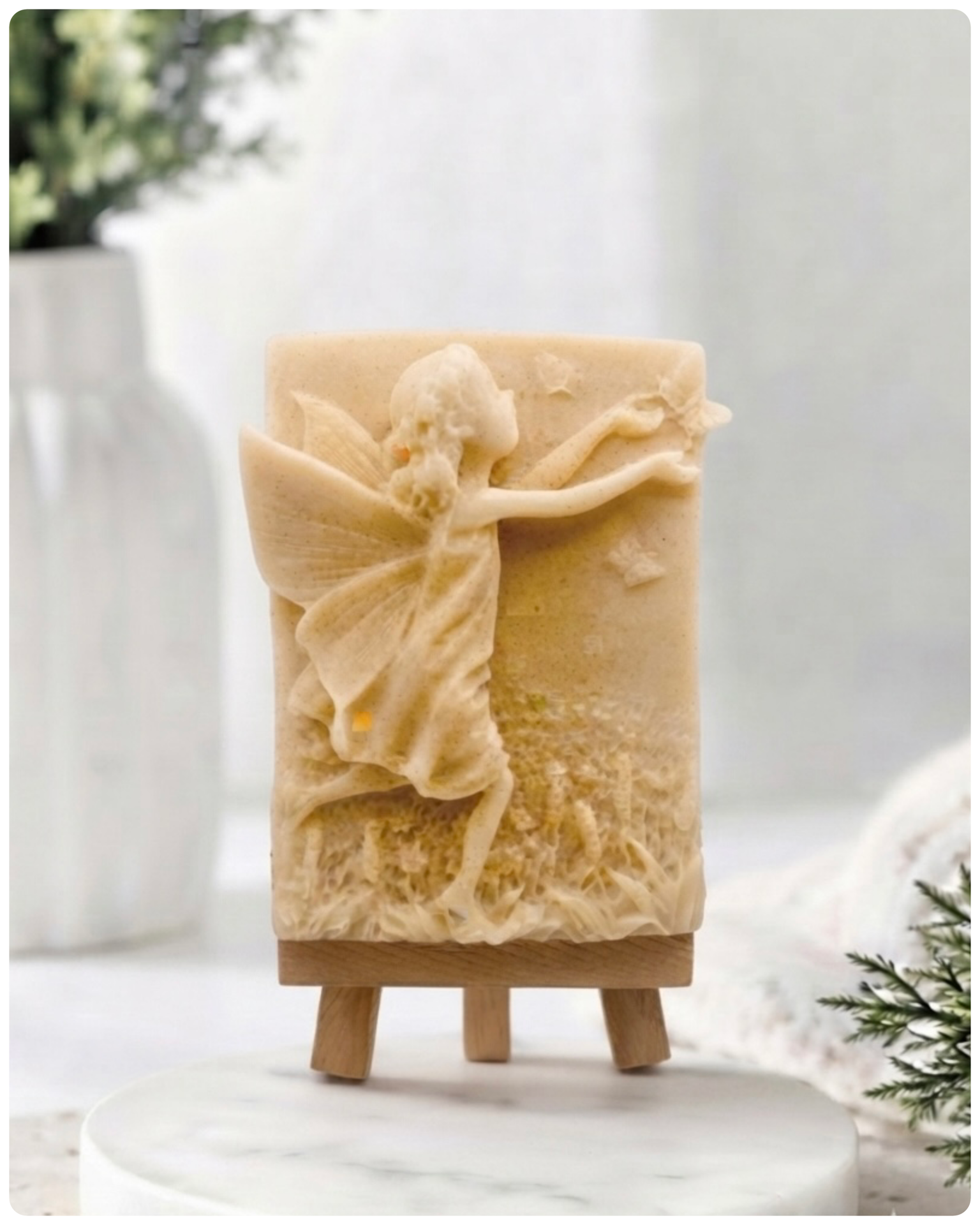 A rectangular handmade vegan soap featuring a detailed relief of a winged fairy reaching towards a butterfly in a meadow of tall grass and flowers, displayed on a miniature wooden easel
