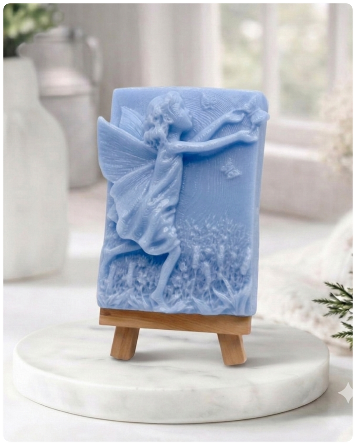 A rectangular handmade vegan soap featuring a detailed relief of a winged fairy reaching towards a butterfly in a meadow of tall grass and flowers, displayed on a miniature wooden easel