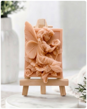 Load image into Gallery viewer, A rectangular handmade vegan soap, featuring a high-relief carving of a seated fairy with large, detailed wings holding a single blossom, displayed on a miniature wooden easel.
