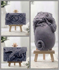 A composite product photo showing a grey sculptural dragon-themed vegan soaps from multiple angles, displayed on small wooden easels against a soft light background. The views include a rectangular plaque with a dragon eye motif, a side view, and a larger rounded form with a curled tail and textured dragon wrapping around it.