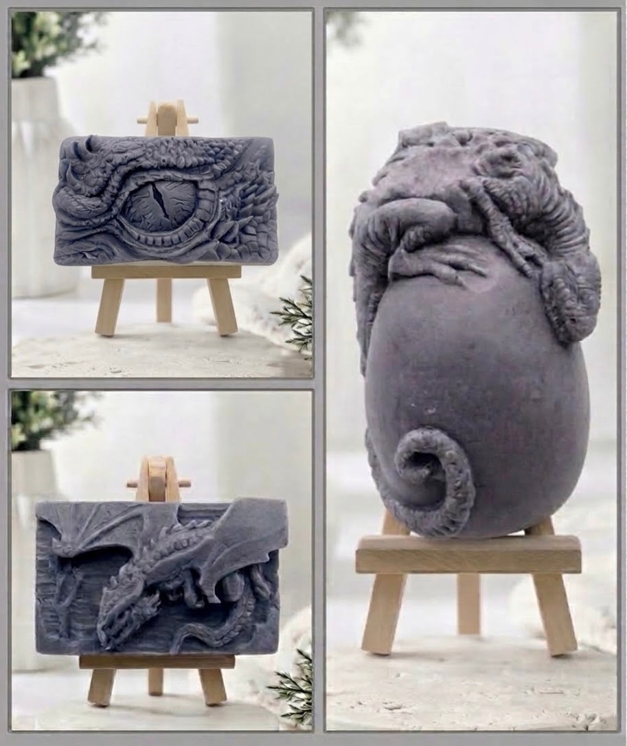 A composite product photo showing a grey sculptural dragon-themed vegan soaps from multiple angles, displayed on small wooden easels against a soft light background. The views include a rectangular plaque with a dragon eye motif, a side view, and a larger rounded form with a curled tail and textured dragon wrapping around it.