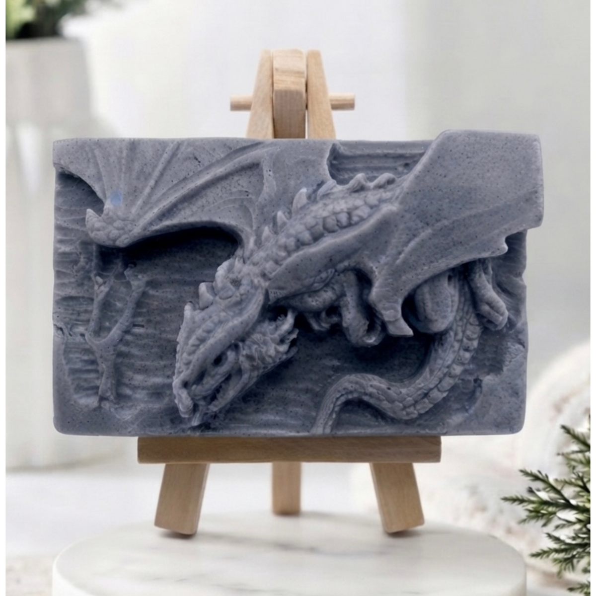 handmade charcoal grey soap bar featuring a detailed relief carving of a dragon, displayed on a small wooden easel in a bright, rustic room with white flowers in the background.