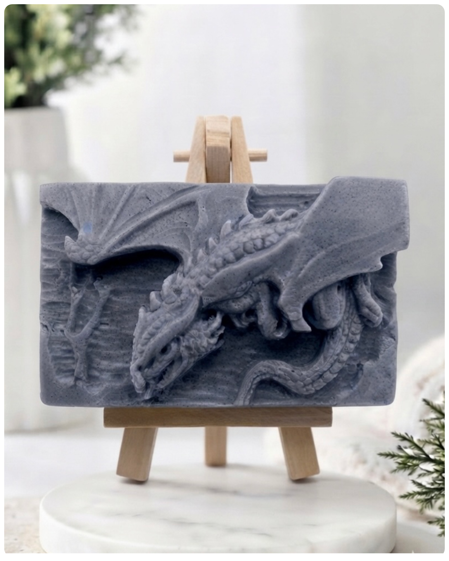 handmade soap bar featuring a detailed 3D relief carving of a dragon, displayed on a small wooden easel in a bright, rustic room with white flowers in the background.