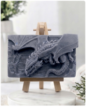 Load image into Gallery viewer, handmade soap bar featuring a detailed 3D relief carving of a dragon, displayed on a small wooden easel in a bright, rustic room with white flowers in the background.

