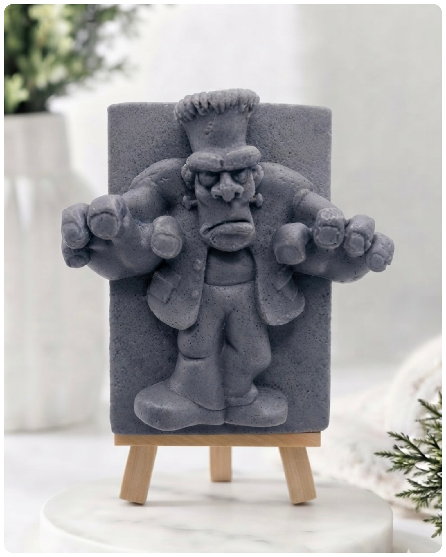 A rectangular handmade vegan soap featuring a high-relief carving of Frankenstein's monster in a classic pose with arms outstretched and a grumpy expression, displayed on a miniature wooden easel.