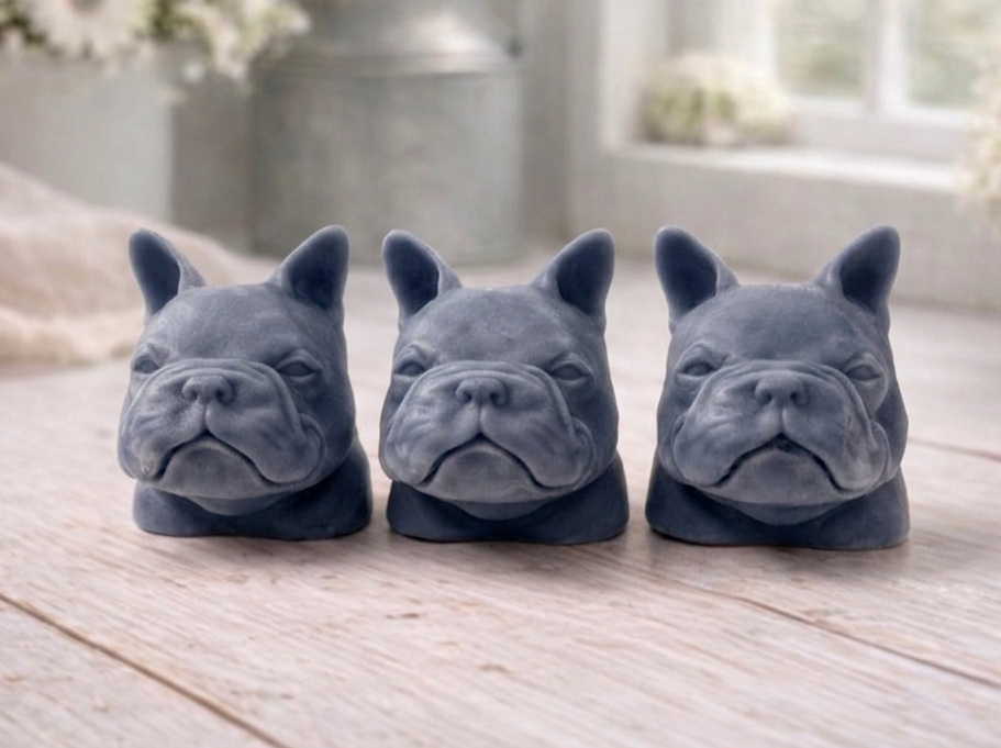 three french bulldog-head shaped bars of soap, side-by-side, on a wooden surface, set in a cosy, rustic bathroom with a vase of white and yellow flowers