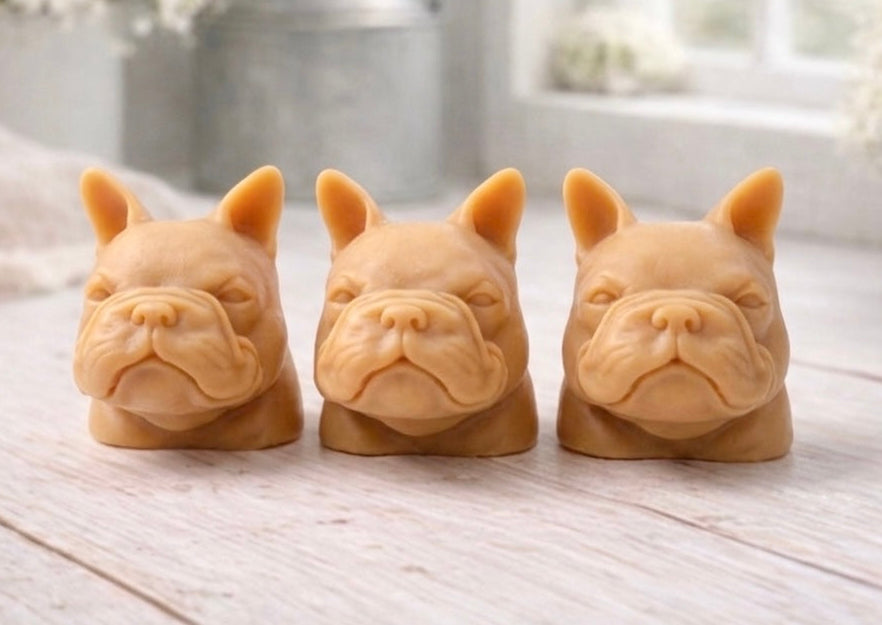 three french bulldog-head shaped bars of soap, side-by-side, on a wooden surface, set in a cozy, rustic bathroom with a vase of white and yellow flowers