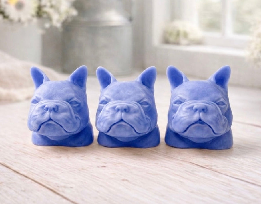 three french bulldog-head shaped bars of soap, side-by-side, on a wooden surface, set in a cozy, rustic bathroom with a vase of white and yellow flowers
