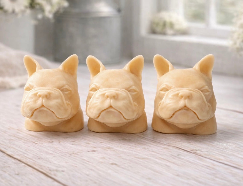 three french bulldog-head shaped bars of soap, side-by-side, on a wooden surface, set in a cozy, rustic bathroom with a vase of white and yellow flowers
