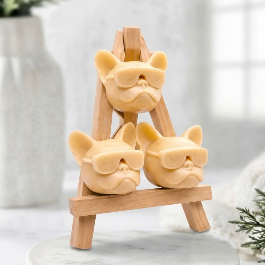 Handcrafted French Bulldog novelty soap gift set. Three artisan, soft yellow coloured Frenchie-head soaps wearing matching sunglasses are stacked and displayed on a natural wooden artist's easel.