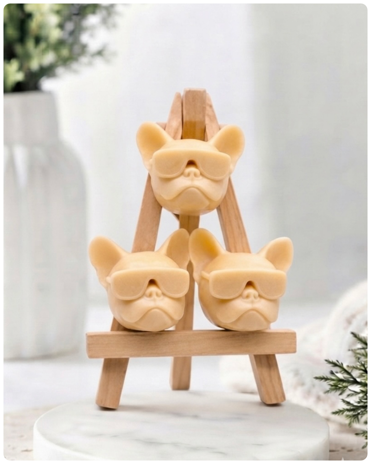 A set of three handmade vegan soaps in the shape of French Bulldog heads wearing sunglasses, displayed on a wooden easel in a bright, modern bathroom setting.