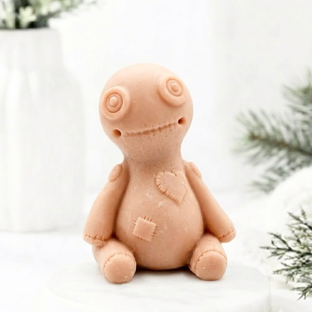 This soft pink voodoo doll soap is highly detailed, including spiral eyes, a stitched mouth, and carved heart and belly patches, against a bright, softly blurred bathroom background
