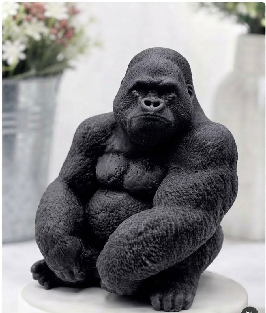 A black gorilla charcoal vegan soap sitting on a white round pedestal, photographed indoors against a softly blurred light background with decorative vases in the distance.