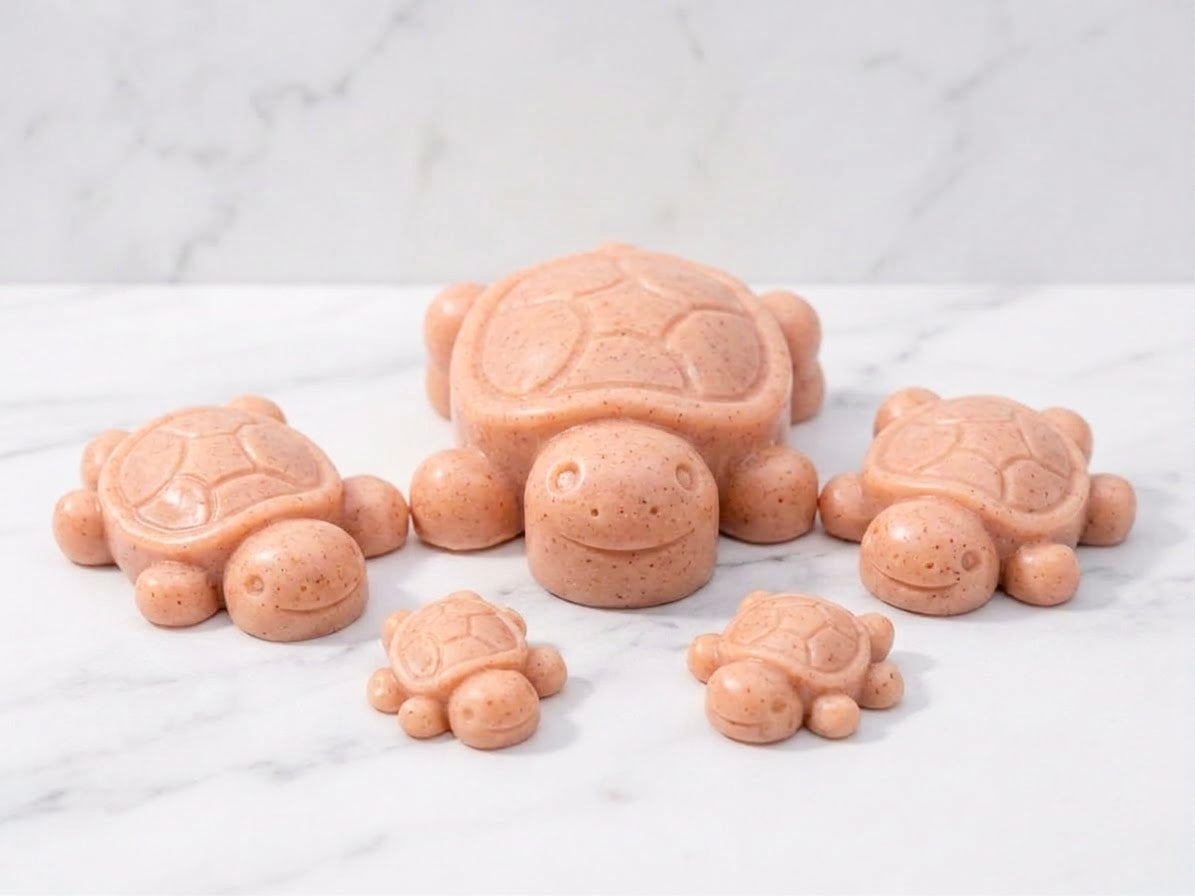 A family-themed cluster of five coral-terracotta coloured handmade turtle soap bars, including one large and two smaller pairs, resting on a white marble countertop. Unique animal soap gift set.