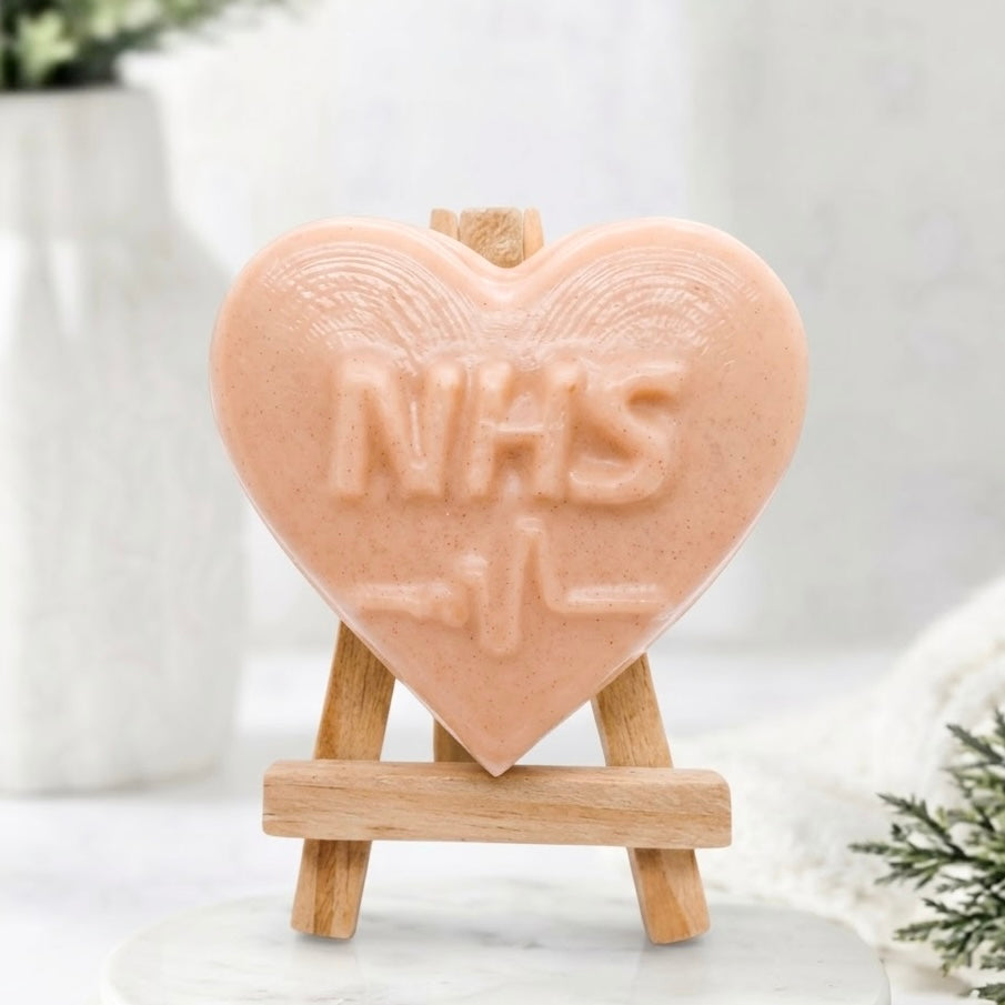 A soft pink, heart-shaped soap with embossed "NHS" lettering and an ECG heartbeat line underneath. The soap stands vertically on a wooden easel resting on a white marble plinth, surrounded by a neutral, soft-focus bathroom display with greenery and a white vase.