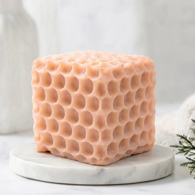 Close-up of a pink, intricate honeycomb-textured vegan soap cube on a white marble coaster in a brightly lit, soft-focus spa setting.
