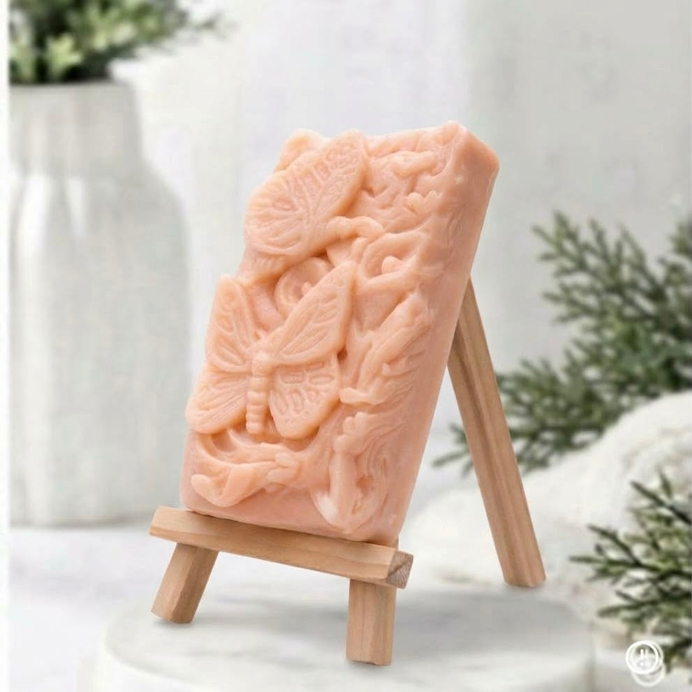 A side view of a delicate pink artisanal soap, standing vertically on a small wooden easel. The soap features a detailed relief carving of a large butterfly with two smaller ones fluttering among swirling floral and leaf motifs. It's displayed on a marble base with a blurred white vase and soft greenery in the background.