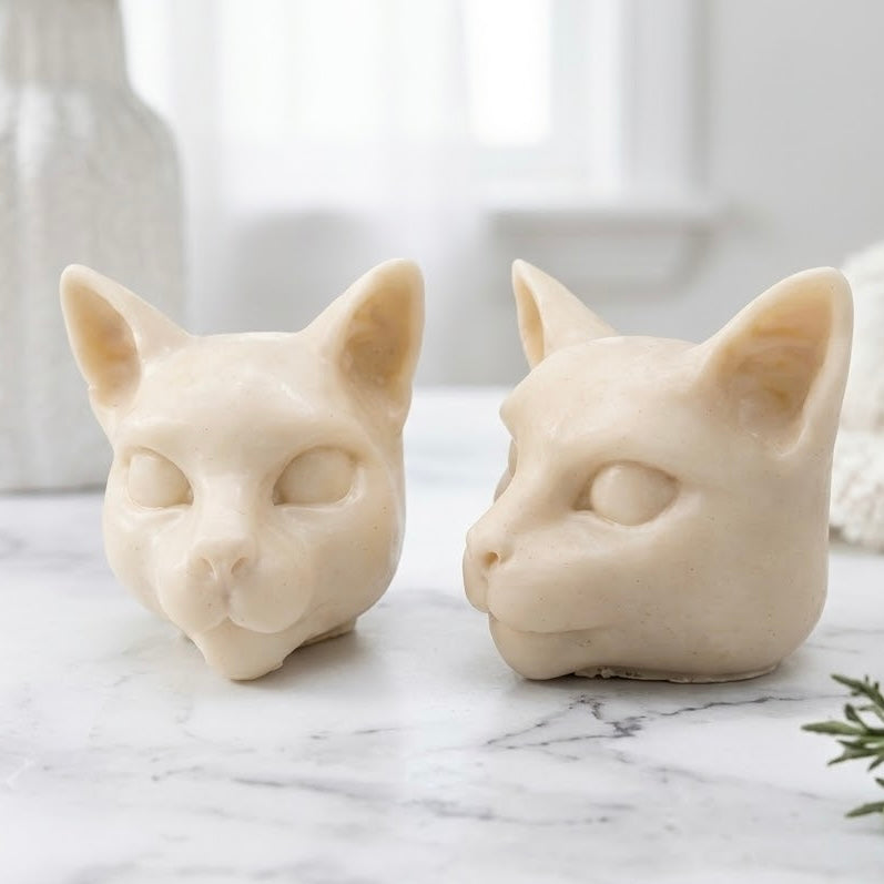 Two detailed, soft yellow vegan soap bars with a smooth matte finish, handmade into Siamese cat heads and resting on a white marble surface.