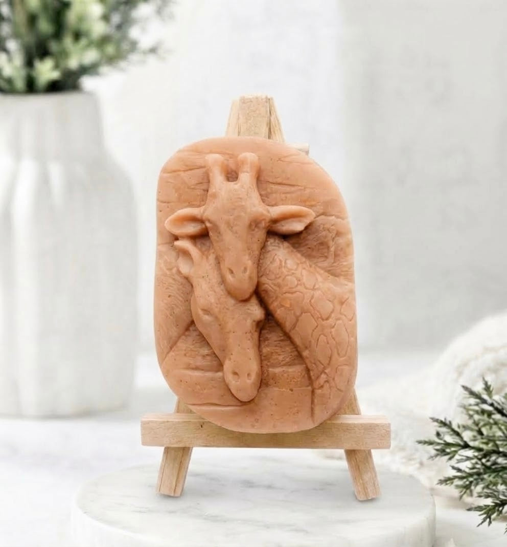 A close-up photograph of a terracotta coloured handmade vegan soap sculpted into two detailed giraffe heads pressed together in a loving embrace. The soap is displayed on a small, light-wood artist's easel, resting on a white marble surface.
