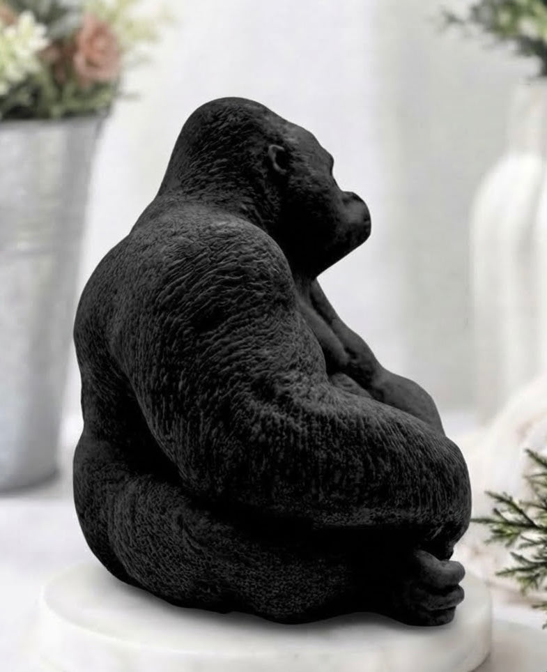 A black gorilla vegan soap sitting cross-legged on a white round pedestal, photographed in a softly blurred indoor setting with flowers and white decor in the background.