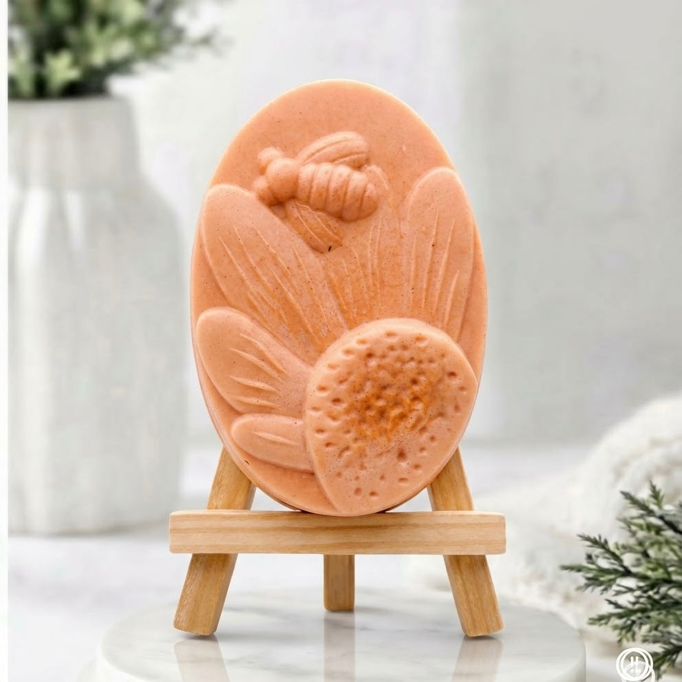 An oval terracotta coloured soap with embossed floral and bee details, displayed on a small wooden stand against a soft, light grey background.