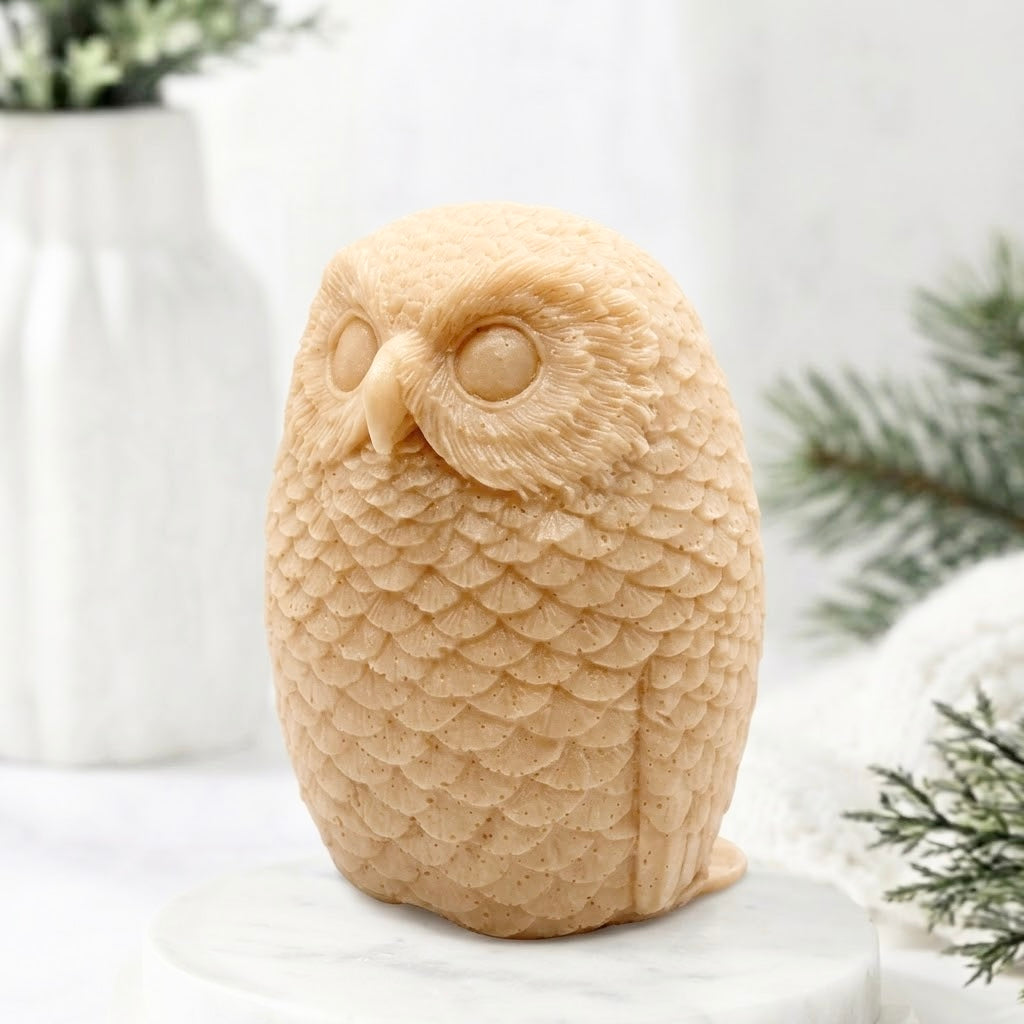 A detailed yellow coloured handmade vegan soap in the shape of an owl with intricate feather textures, displayed on a light wooden table with white flowers in the background.