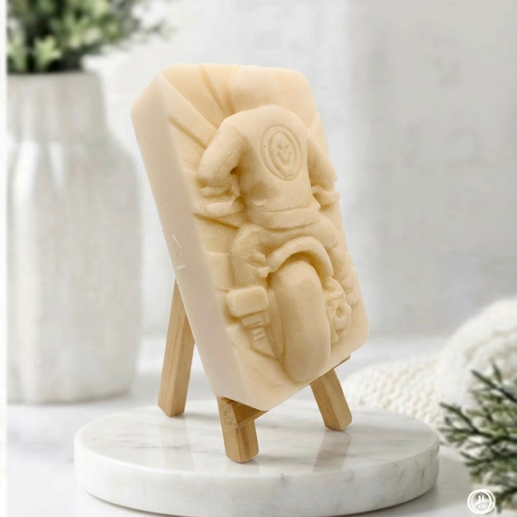 A side view of a yellow-coloured natural handmade motorbike soap bar with carved rider and skull patch design, displayed on a wooden easel stand