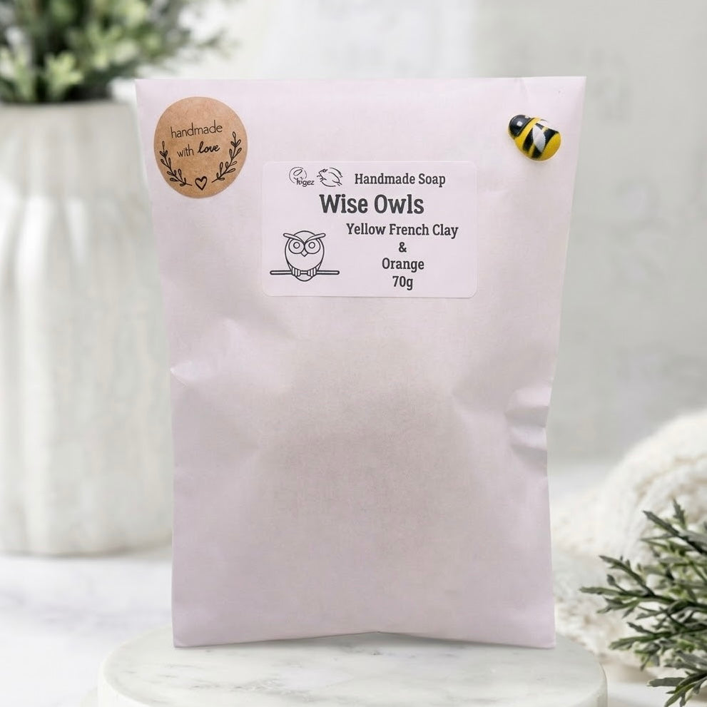 A 70g bag of Wise Owls Yellow French Clay & Orange handmade soap by Paper Owl, displayed on a white marble pedestal with a soft-focus background.
