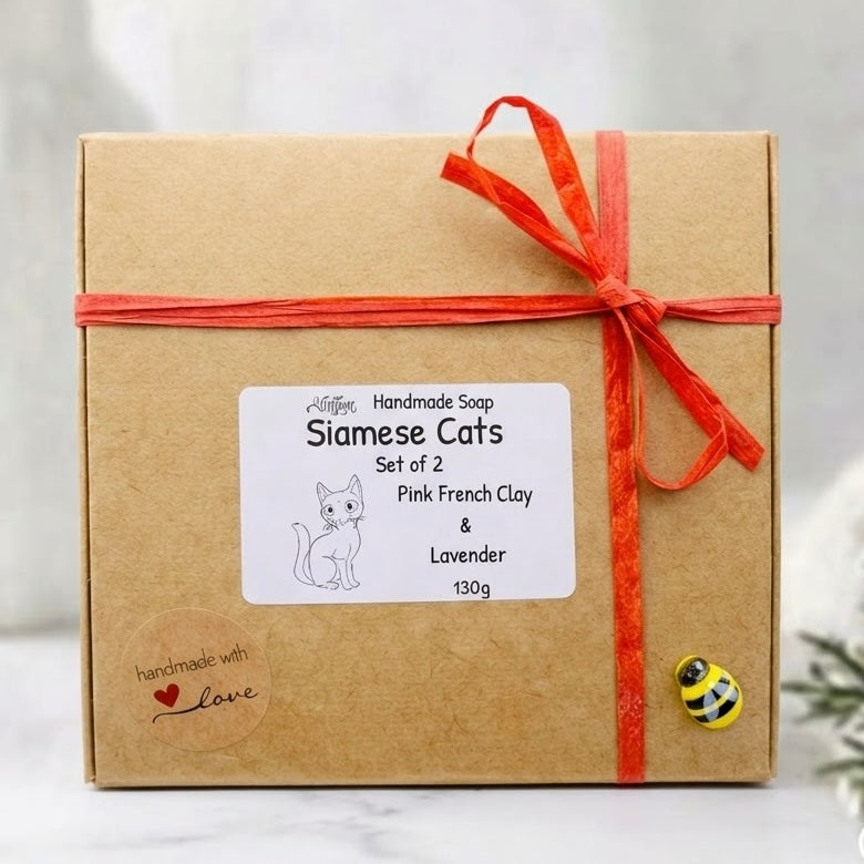A closed square brown kraft paper gift box tied with a red raffia ribbon. A central white label features a cat drawing and text reading: "Handmade Soap, Siamese Cats, Set of 2, Pink French Clay & Lavender, 130g." A tiny bee charm and a 'handmade with love' sticker are also on the box.