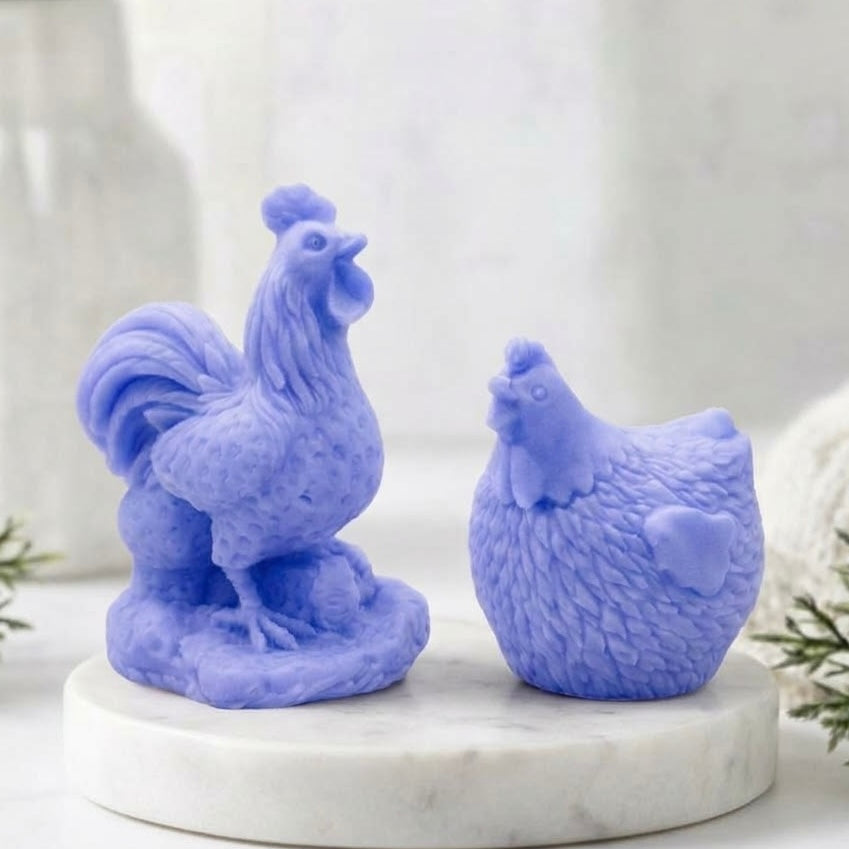 Side profile of two beautifully detailed, vibrant blue, handmade vegan soap bars - one cockerel and one hen - sitting on a white marble coaster against a bokeh background of a window and blurry greenery.