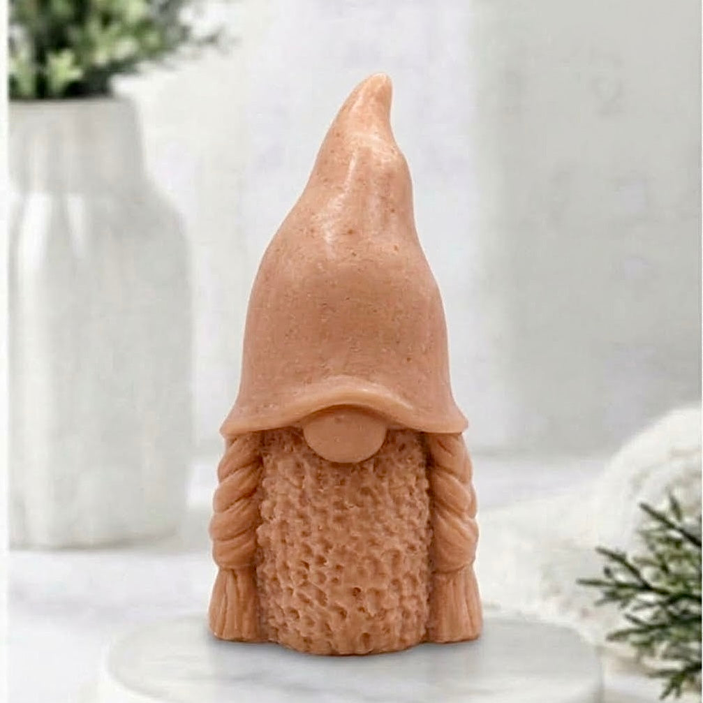 A handmade terracotta coloured vegan soap in the shape of a female gnome (or gonk), featuring a tall, slightly curved hat pulled over the eyes and two thick, textured plaits framing a textured dress.
