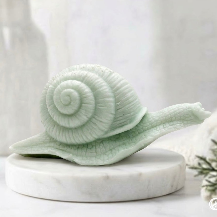 A highly detailed handmade vegan soap in a mint green colour, sculpted in the shape of a realistic snail with a prominent spiralled shell and a textured, leathery body, displayed on a light wooden surface.
