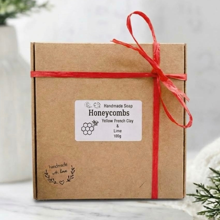 A square brown kraft gift box tied with a red ribbon and a white label reading 'Handmade Soap Honeycombs Yellow French Clay & Lime 100g', photographed on a light marble-like surface with soft decorative greenery in the background.