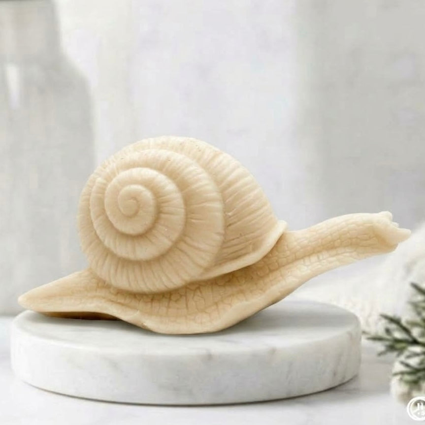 A highly detailed handmade vegan soap in a soft yellow colour, sculpted in the shape of a realistic snail with a prominent spiralled shell and a textured, leathery body, displayed on a light wooden surface.