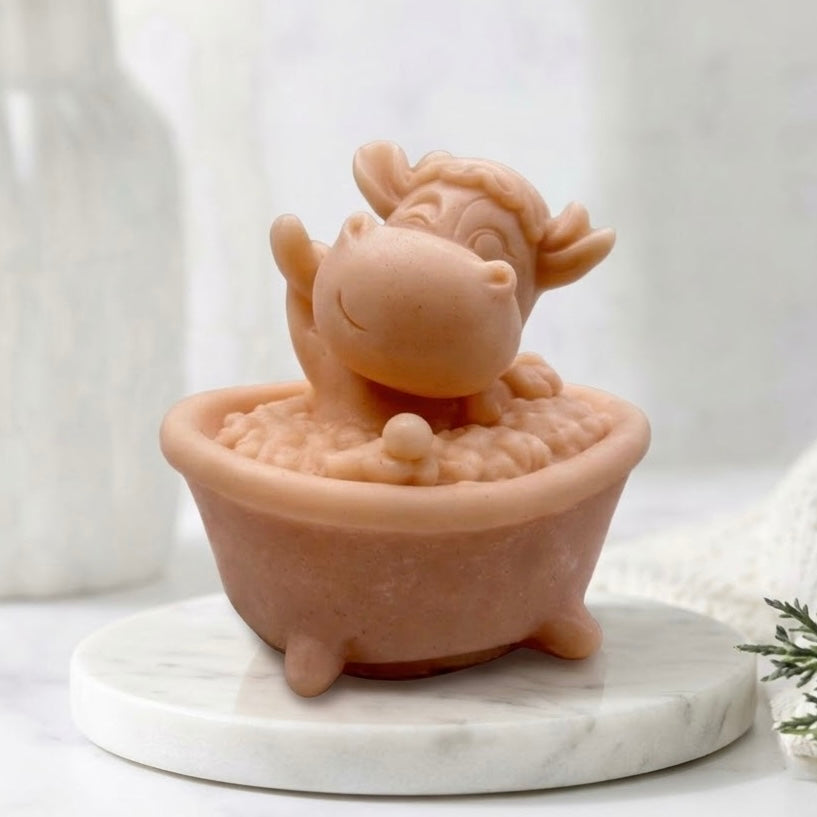 A terracotta coloured novelty vegan soap bar in a cute cartoon cow waving happily while sitting in a miniature footed bathtub filled with soap bubbles.