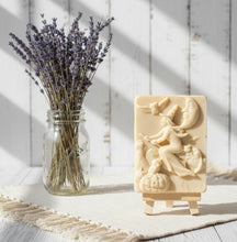 Load image into Gallery viewer, A beautiful handmade vegan soap featuring a detailed image of a witch flying on a broomstick past a crescent moon and owls, displayed on a wooden easel against a whimsical farmhouse decor background.
