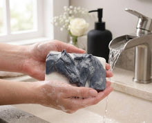 Load image into Gallery viewer, A rectangular handmade vegan soap featuring a detailed relief of two horse heads leaning affectionately towards each other, finished in a slate-grey tone against a white soap base and displayed on a miniature wooden easel.
