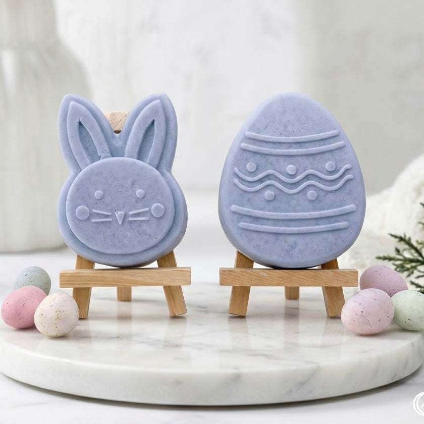 Luxury Easter soap gift set with two artisan vegan soaps on mini wooden easels. Features a detailed periwinkle-blue bunny face soap and a matching patterned Easter egg soap, set on a round marble tray with speckled pastel miniature eggs for decoration
